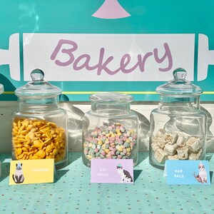 Puede incluir: Tres tarros de vidrio llenos de golosinas para gatos, etiquetados como "Goldfish", "Cat Treats" y "Hair Balls". Los tarros est&aacute;n sobre una mesa con un mantel de lunares azules y blancos. Los tarros est&aacute;n frente a un letrero que dice "Bakery" con un gr&aacute;fico de rodillo.