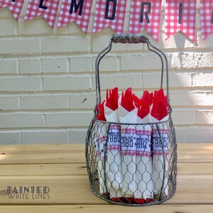 May include: A wire basket filled with red plastic utensils wrapped in white napkins with a red and white checkered pattern and the text "Memorial Day BBQ" printed on them. The basket is sitting on a wooden table.