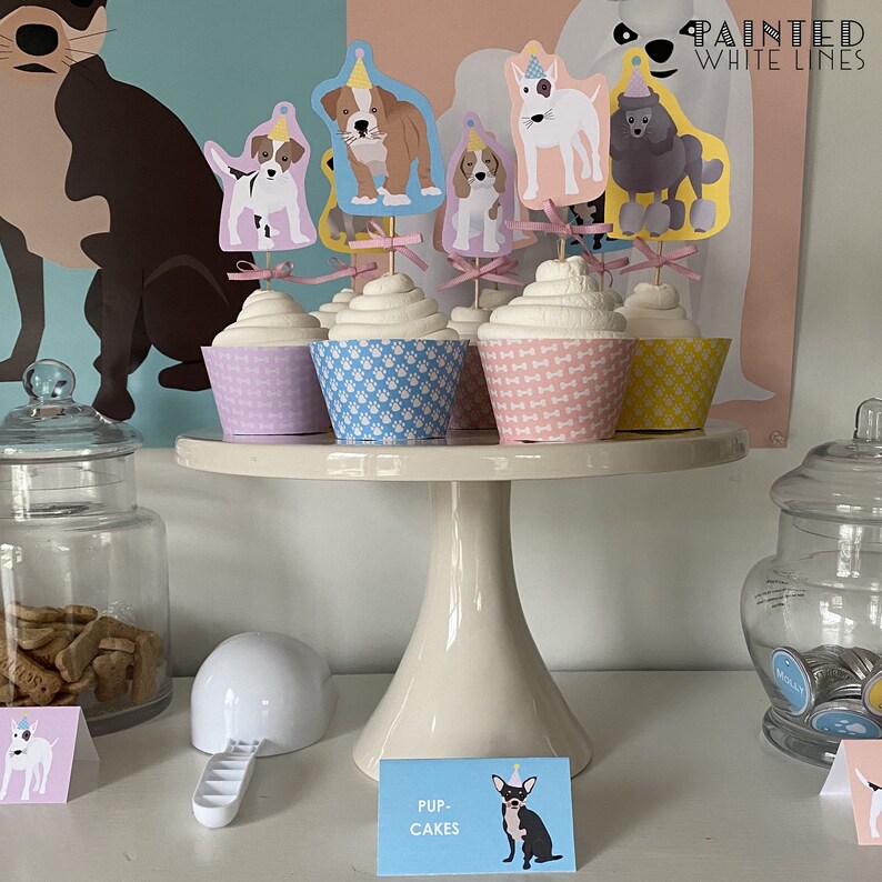 May include: A white cake stand with a tier holds six cupcakes decorated with dog-themed toppers. The cupcakes are in pink, blue, and yellow wrappers. The stand is on a white surface with a sign that reads "Pup-Cakes".