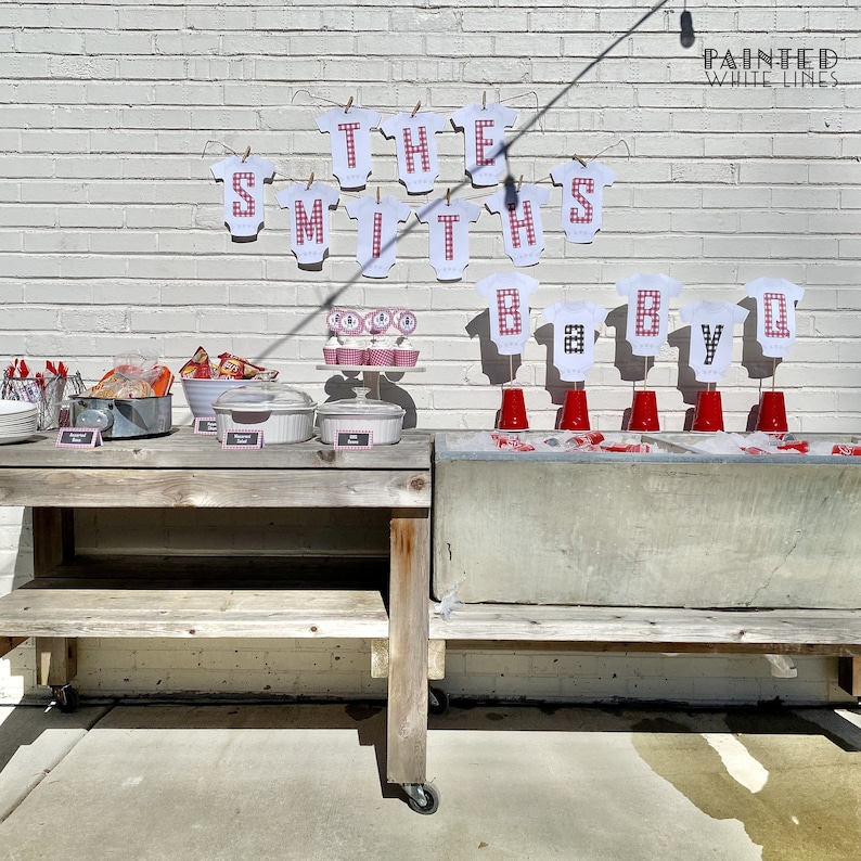 May include: A baby shower party setup with a wooden table and a metal cooler. The table is decorated with red and white gingham paper and has a sign that says "The Smiths Baby BBQ".