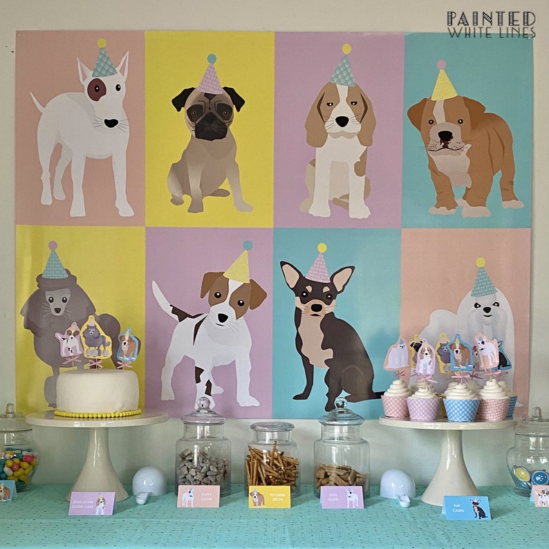 May include: A colorful party backdrop with illustrations of dogs wearing party hats. The backdrop is decorated with a yellow, pink, and blue color scheme. The backdrop is hung on a wall with a table in front of it. The table is decorated with a cake, jars of treats, and cupcakes. The cupcakes are decorated with dog-themed toppers. The text "PAINTED WHITE LINES" is at the top of the backdrop.