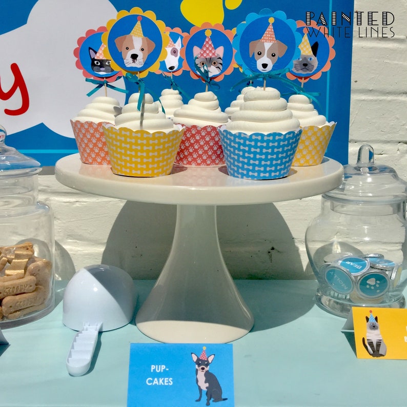 Pu&ograve; includere: Un porta torte bianco con sei cupcake decorati con topper a forma di cane e gatto. I cupcake sono ricoperti di glassa blu, gialla e rosa. Il porta torte &egrave; su una superficie blu chiaro con un cartello che dice "Pup Cakes".