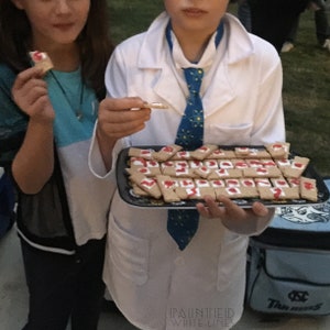 May include: A tray of decorated cookies with red icing. The cookies are arranged in rows on a black tray with a gold design. A person wearing a white lab coat and a blue tie is holding the tray.
