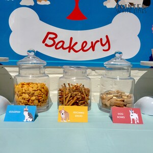 Pu&ograve; includere: Tre barattoli di vetro pieni di dolcetti per cani, etichettati "Goldfish", "Fetching Sticks" e "Dog Bones". I barattoli sono su un tavolo blu chiaro con un cartello sopra che dice "Bakery".