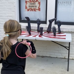 May include: A girl throws a ring at a game with a red and white striped target. The target has several black, severed hands and feet sticking out of it. The sign above the game reads "Dead Ringer".