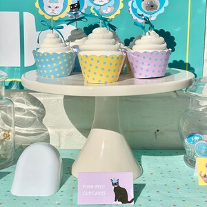 May include: A white cake stand with three cupcakes decorated with cat faces wearing party hats. The cupcakes are topped with yellow and blue polka dot cupcake liners. A sign reads "Purr-fect Cupcakes" with a black cat wearing a party hat.