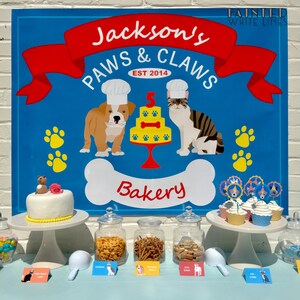 Pu&ograve; includere: Un cartello blu e rosso con il testo "Jackson's Paws & Claws Bakery Est 2014" con un cane e un gatto dei cartoni animati che indossano cappelli da chef. Ci sono cupcake con decorazioni di cane e gatto su un porta torte bianco. Ci sono barattoli di dolcetti per cani su un tavolo con etichette che dicono "Gourmet Biscuits", "Dog Bones", "Chicken Bites" e "Pup Cakes".