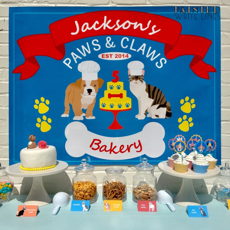Pu&ograve; includere: Un cartello blu e rosso con il testo "Jackson's Paws & Claws Bakery Est 2014" con un cane e un gatto dei cartoni animati che indossano cappelli da chef. Ci sono cupcake con decorazioni di cane e gatto su un porta torte bianco. Ci sono barattoli di dolcetti per cani su un tavolo con etichette che dicono "Gourmet Biscuits", "Dog Bones", "Chicken Bites" e "Pup Cakes".