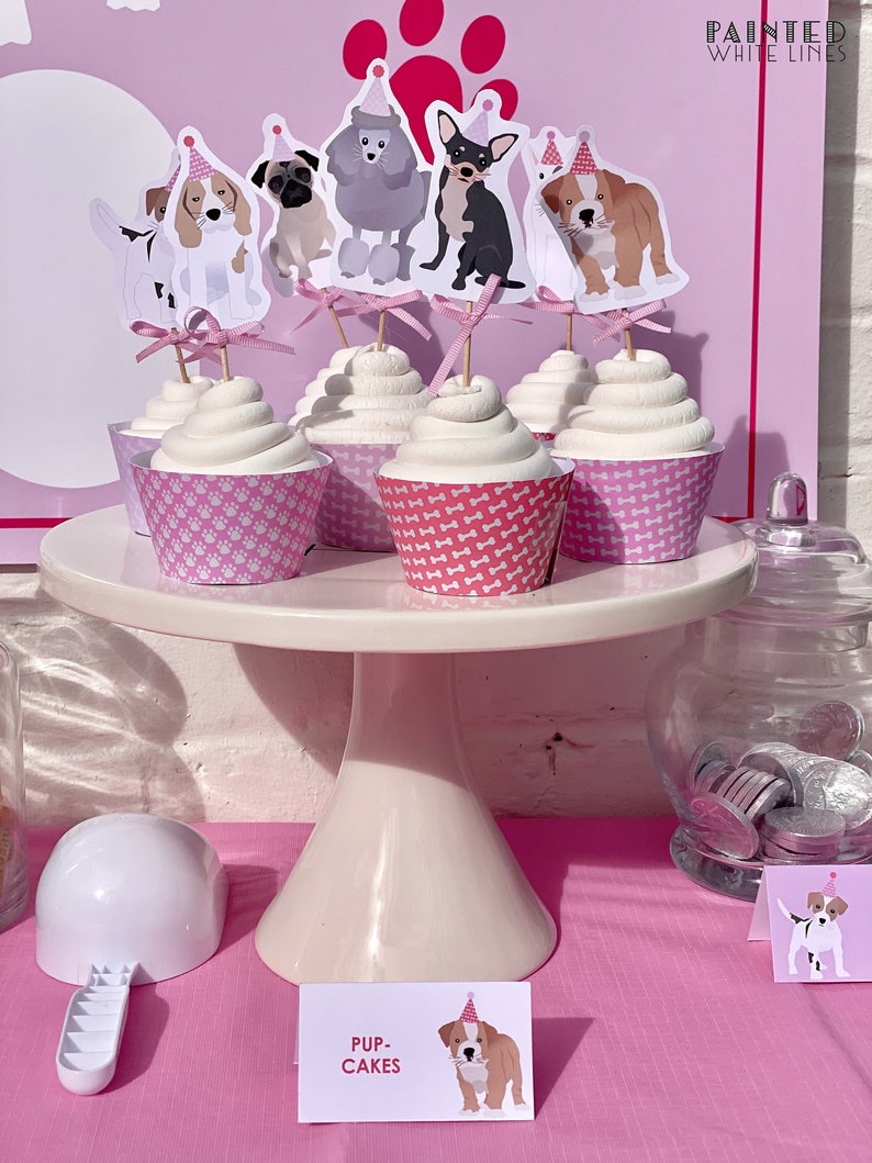 Może przedstawiać: R&oacute;żowo-biały stojak na babeczki z babeczkami ozdobionymi topperami w kształcie ps&oacute;w. Babeczki są oznaczone jako "Pup-Cakes".
