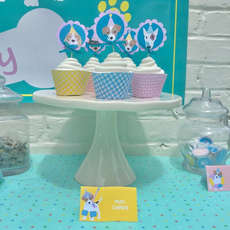 May include: A white cake stand with four cupcakes decorated with dog-themed toppers. The cupcakes are yellow, blue, and pink. The stand is on a turquoise table with white polka dots. A sign reads "Pup Cakes".