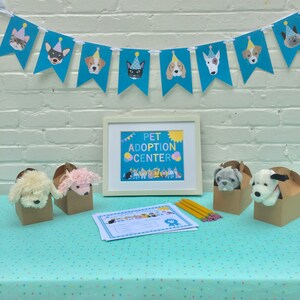 May include: A pet adoption center display featuring a banner with dog and cat illustrations wearing party hats. The framed sign reads "Pet Adoption Center." Plush toy dogs are in cardboard boxes. Pencils and adoption certificates are also on display.