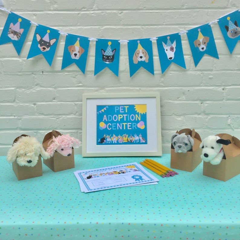 May include: A pet adoption center display featuring a banner with dog and cat illustrations wearing party hats. The framed sign reads "Pet Adoption Center." Plush toy dogs are in cardboard boxes. Pencils and adoption certificates are also on display.