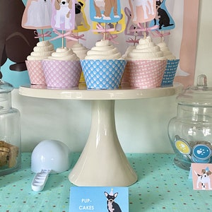 May include: A table with a white cake stand holding cupcakes decorated with dog-themed toppers. The cupcakes are on a blue and white polka dot tablecloth. The sign reads "Pup-Cakes".