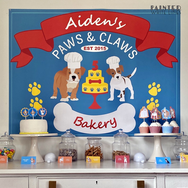 May include: A blue and red party backdrop with the text "Aiden's Paws & Claws Est 2015 Bakery". Two cartoon dogs wearing chef hats are on either side of a cake with a number 5 on top. There are cupcakes with dog toppers on a white cake stand.