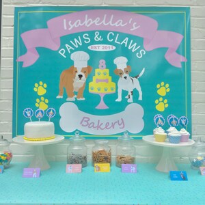 May include: A turquoise and pink banner with the text "Isabella's Paws & Claws Est 2011 Bakery" with a bone-shaped graphic. A table with a white cake decorated with dog-themed toppers, jars of treats, and cupcakes with dog-themed toppers.