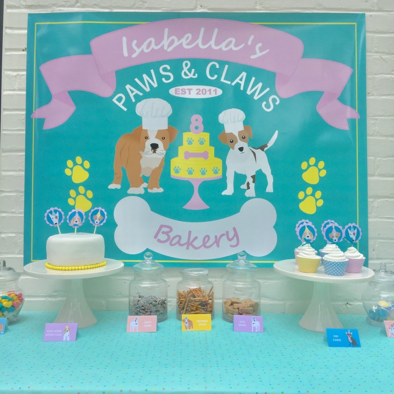 May include: A turquoise and pink banner with the text "Isabella's Paws & Claws Est 2011 Bakery" with a bone-shaped graphic. A table with a white cake decorated with dog-themed toppers, jars of treats, and cupcakes with dog-themed toppers.