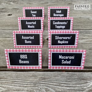 May include: Eight red and white gingham table cards with black text. The cards list various food and beverage options for a party or event. The text on the cards reads: Sweet Tea, Assorted Meats, Assorted Buns, BBQ Beans, Adult Beverages, Condiments/Toppings, Silverware/Napkins, Macaroni Salad.