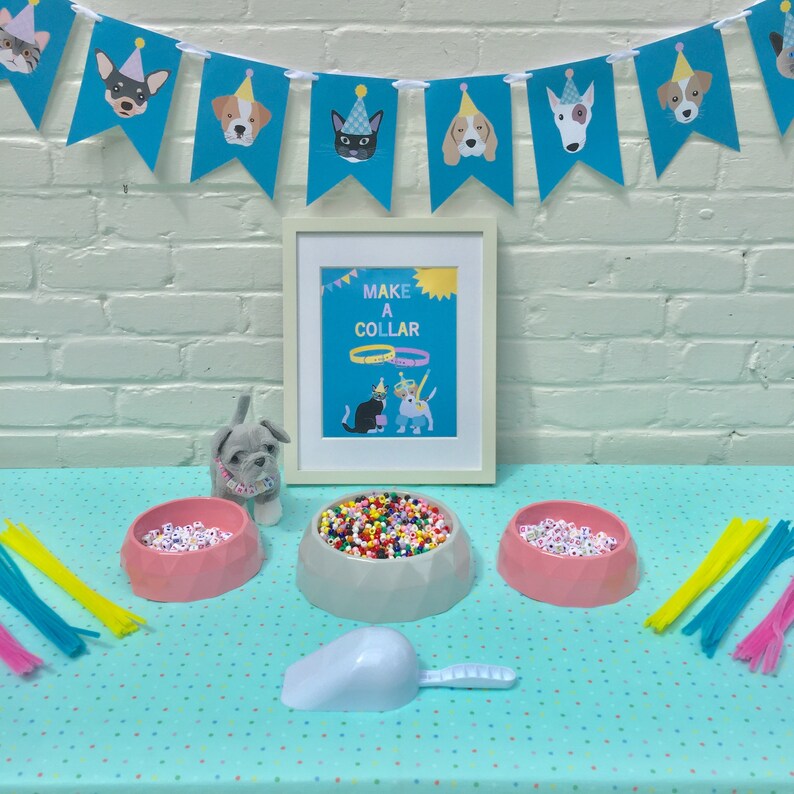 May include: A party-themed craft kit with a blue banner featuring dog faces wearing party hats. The kit includes a framed sign that says "MAKE A COLLAR", three bowls of beads, pipe cleaners, a scoop, and a stuffed dog.