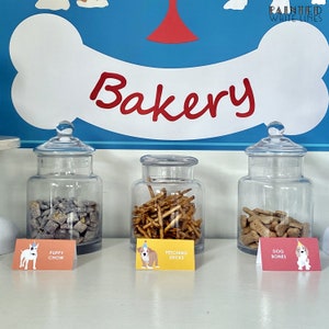 May include: Three glass jars filled with dog treats, labeled "Puppy Chow", "Fetching Sticks", and "Dog Bones". The jars are on a white counter with a blue and white dog bone sign above them that says "Bakery".