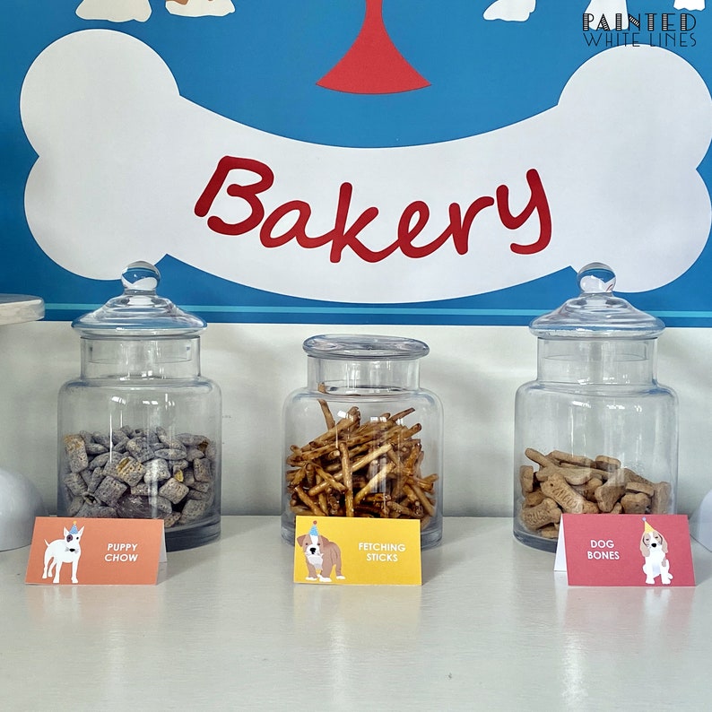 May include: Three glass jars filled with dog treats, labeled "Puppy Chow", "Fetching Sticks", and "Dog Bones". The jars are on a white counter with a blue and white dog bone sign above them that says "Bakery".
