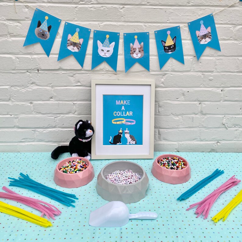 May include: A blue and white banner with cats wearing party hats reads "Make a Collar".  A black cat plush toy sits in front of a framed print with the same banner design.  There are bowls of colorful beads and pipe cleaners on a table with a polka dot tablecloth.