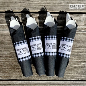 May include: Four black napkins with white and black checkered paper rings that say "Kristy & Mike's I Do BBQ" with a ring icon. The napkins are wrapped around white plastic utensils.