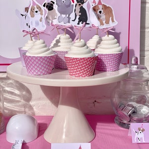 May include: A pink and white cupcake stand with cupcakes decorated with dog toppers. The cupcakes are labeled "Pup-Cakes".