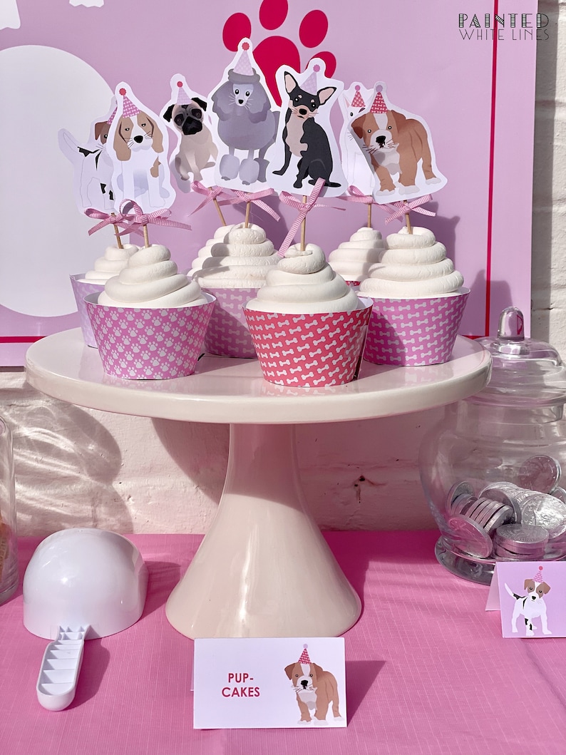 May include: A pink and white cupcake stand with cupcakes decorated with dog toppers. The cupcakes are labeled "Pup-Cakes".