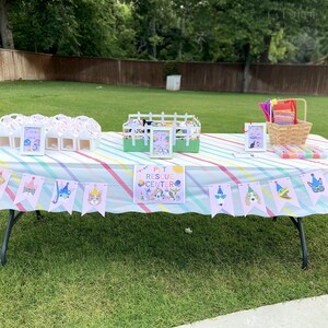 May include: A table with a rainbow striped tablecloth set up outdoors for a party. There are party favours in white boxes, a sign that says "Pet Rescue Center", and a banner with cartoon animals wearing party hats.