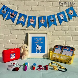 May include: A blue and white banner with cartoon dogs wearing party hats hangs above a table with a red toy doctor's bag, a white stuffed dog, a framed print that says "Give your pet a check-up", two clipboards, a yellow basket, and a toy stethoscope.