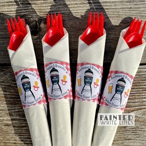 May include: Four white napkins with red and white checkered paper bands wrapped around them. The bands have a design with a grill, beer, and a pacifier with the text "Barbeque & Brewskis" and "The Smiths" printed on them. The napkins are wrapped around red plastic forks and spoons.