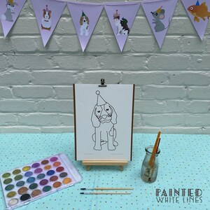 May include: A black and white line drawing of a dog wearing a party hat on an easel. The dog is surrounded by a set of watercolors, paintbrushes, and a jar of water. The background is a white brick wall with a purple and white bunting.
