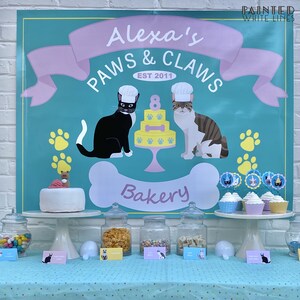 Puede incluir: Un banner de color turquesa y rosa con el texto "Alexa's Paws & Claws Bakery Est 2011" con un gato blanco y negro y un gato atigrado con gorros de chef. El banner est&aacute; decorado con huellas de patas y un hueso. Hay cupcakes con adornos de gato en un soporte de pastel blanco.