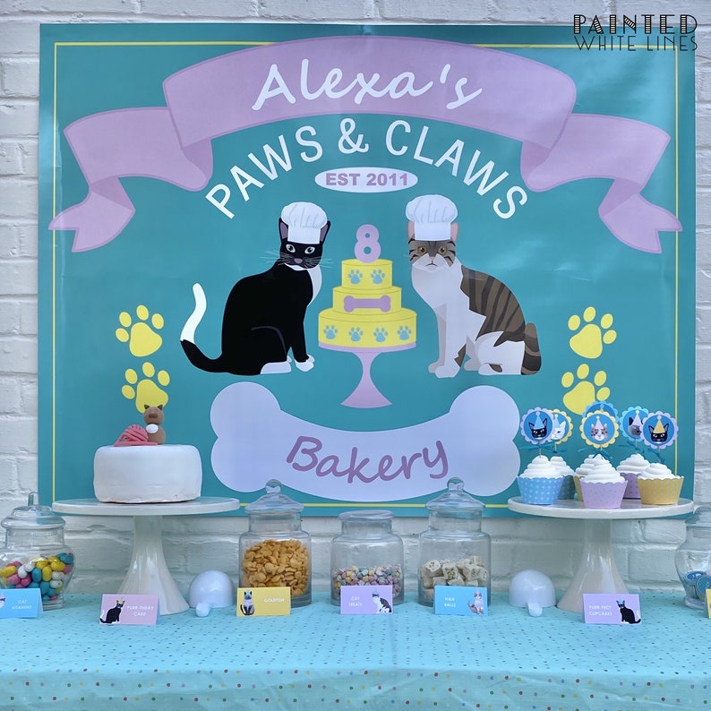 Puede incluir: Un banner de color turquesa y rosa con el texto "Alexa's Paws & Claws Bakery Est 2011" con un gato blanco y negro y un gato atigrado con gorros de chef. El banner est&aacute; decorado con huellas de patas y un hueso. Hay cupcakes con adornos de gato en un soporte de pastel blanco.