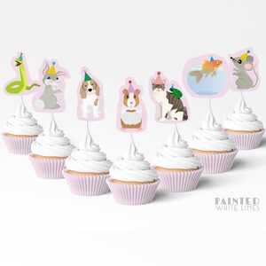 May include: Seven cupcakes with pink liners topped with white frosting. Each cupcake has a different animal wearing a party hat on a toothpick: a snake, a rabbit, a dog, a guinea pig, a cat, a goldfish in a bowl, and a mouse.
