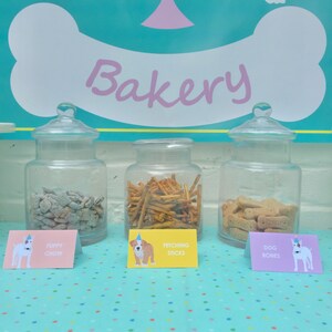 May include: Three glass jars filled with dog treats, labeled "Puppy Chow", "Fetching Sticks", and "Dog Bones". The jars are on a table with a turquoise and white polka dot tablecloth. The sign above the jars reads "Bakery".