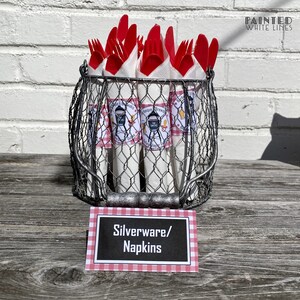 Op de afbeelding: Een draadmand gevuld met rood plastic bestek en witte servetten die zijn ingepakt met barbecue-thema labels. De mand staat op een houten tafel met een bord waarop "Besteck/Servetten" staat.