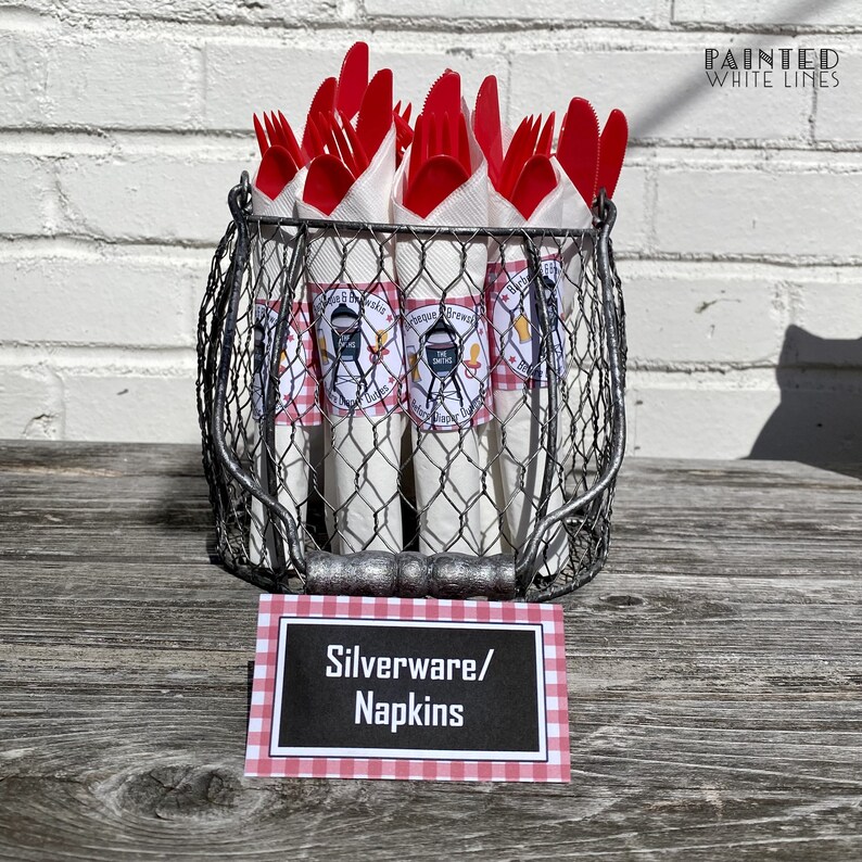 Op de afbeelding: Een draadmand gevuld met rood plastic bestek en witte servetten die zijn ingepakt met barbecue-thema labels. De mand staat op een houten tafel met een bord waarop "Besteck/Servetten" staat.