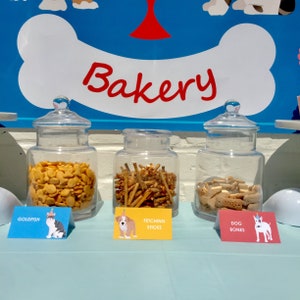 May include: Three glass jars filled with dog treats, labeled "Goldfish", "Fetching Sticks", and "Dog Bones". The jars are on a light blue table cloth with a sign above that says "Bakery".