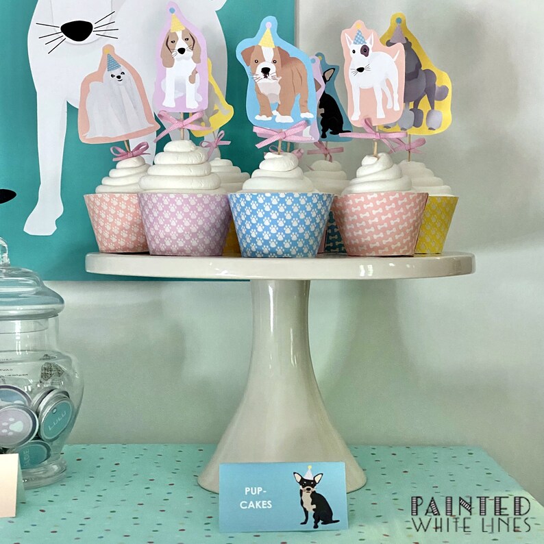 Op de afbeelding: Een witte taartstandaard met zes cupcakes versierd met hondenthema toppers. De cupcakes zijn in roze, blauwe en gele verpakkingen. De standaard staat op een turquoise tafel met witte stippen. Een bordje staat "Pup-Cakes".