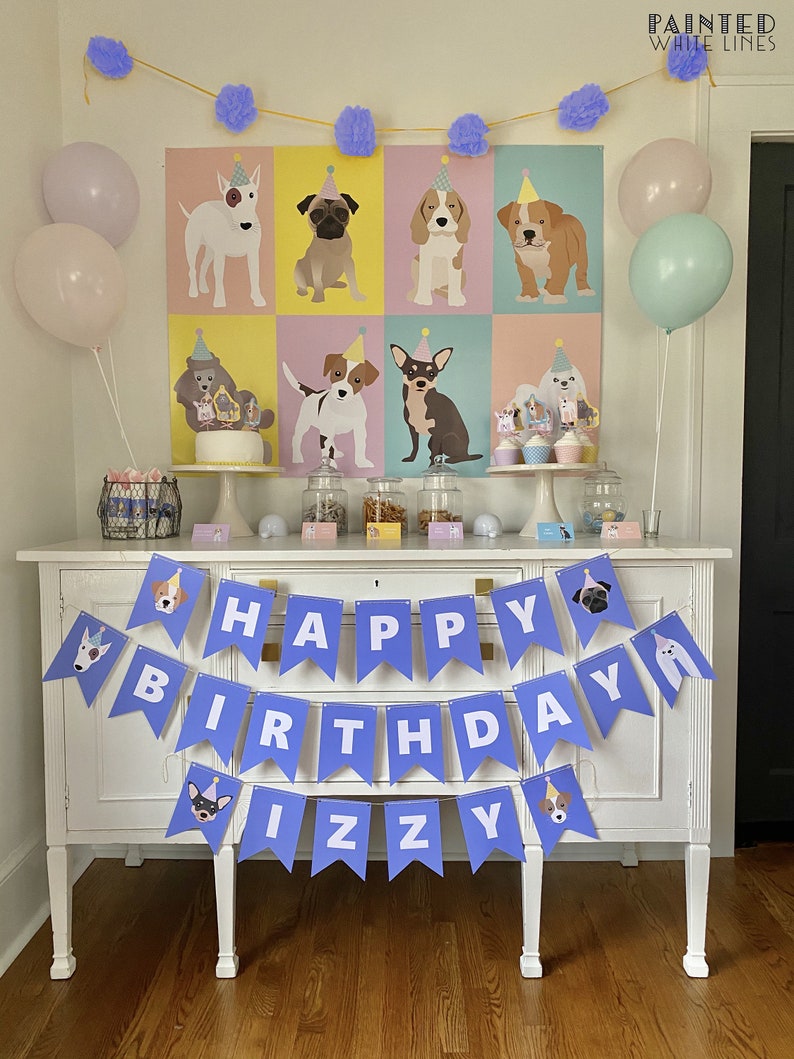 Puede incluir: Una pancarta morada y blanca 'Feliz Cumplea&ntilde;os Izzy' con ilustraciones de perros cuelga sobre una mesa de buffet blanca decorada para una fiesta con tem&aacute;tica de perros. La mesa est&aacute; preparada con un pastel, cupcakes y otras golosinas. Hay globos y un p&oacute;ster con ilustraciones de perros en el fondo.