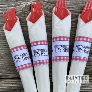 May include: Four white napkins with red and white gingham paper rings. The rings have the text "Kristy & Mike's I Do BBQ" printed on them. The napkins are wrapped around red plastic forks and spoons.