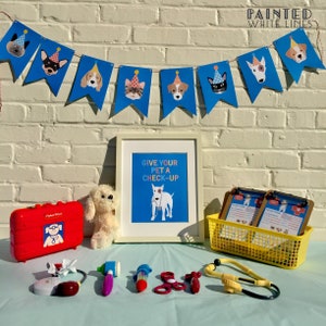 May include: A blue and white banner with cartoon dogs wearing party hats hangs above a table with a red toy doctor's bag, a white stuffed dog, a framed print that says "Give your pet a check-up", two clipboards, a yellow basket, and a toy stethoscope.