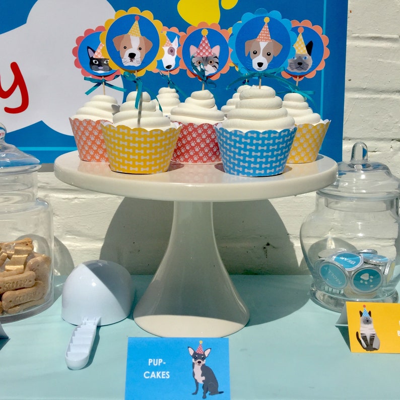 May include: A white cake stand with six cupcakes decorated with dog and cat toppers. The cupcakes are in blue, yellow, and pink wrappers. The cupcakes are on a white cake stand with a blue background. The sign says "Pup Cakes".