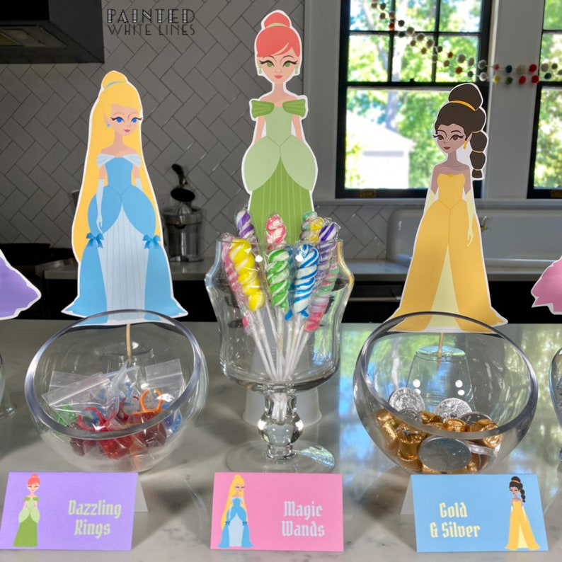 May include: A princess-themed party favor table with three glass bowls filled with candy and small toys. The bowls are labeled "Dazzling Rings", "Magic Wands", and "Gold & Silver".  There are princess character cutouts on sticks in front of each bowl.