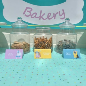 May include: Three glass jars filled with dog treats labeled "Dog Bones", "Fetching Sticks", and "Puppy Chow". The jars are on a blue and white polka dot table cloth with the word "Bakery" in pink letters above them.