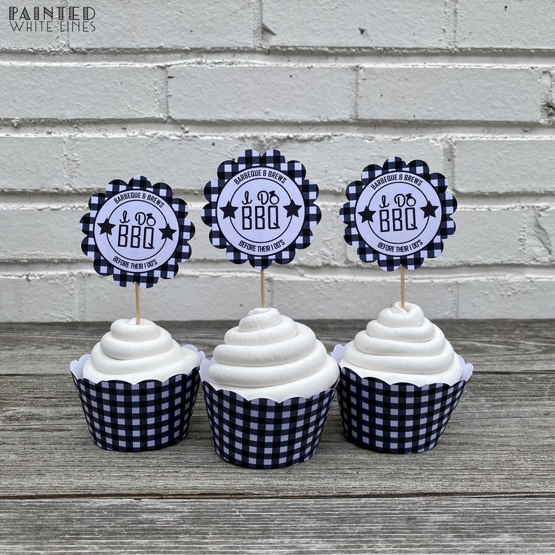 May include: Three white cupcakes with black and white checkered cupcake liners. Each cupcake has a round, scalloped, black and white checkered paper topper with the text "I Do BBQ" and "Barbeque & Brews" in white.