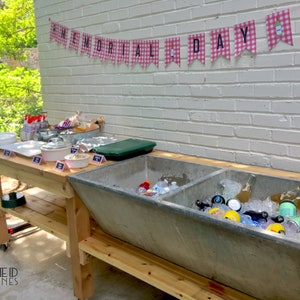 May include: A wooden table with a large, gray concrete trough used as a cooler. The trough is filled with ice and beverages. The table is decorated with a red and white checkered banner that reads "MEMORIAL DAY".