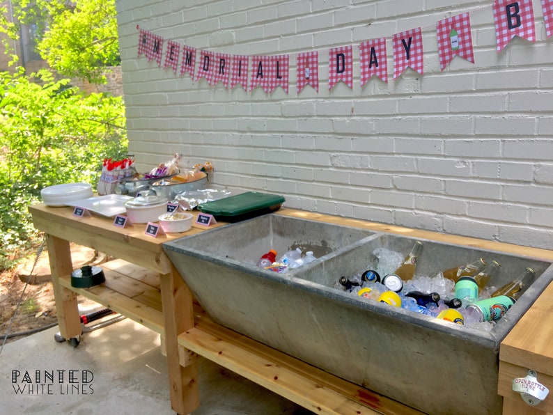 May include: A wooden table with a large, gray concrete trough used as a cooler. The trough is filled with ice and beverages. The table is decorated with a red and white checkered banner that reads "MEMORIAL DAY".
