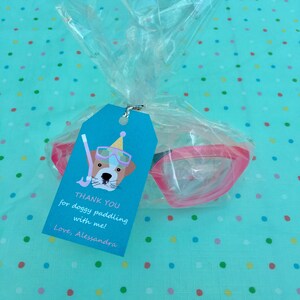 May include: A blue and white thank you tag with a cartoon dog wearing a party hat and goggles. The tag says "Thank you for doggy paddling with me! Love, Alessandra". A pink pair of swimming goggles are in a clear plastic bag.